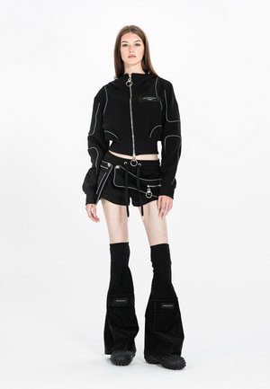 Joven mujer de pie con una chaqueta negra corta con cremallera, minifalda con detalles de cremallera y calentadores de piernas negros y holgados sobre zapatos gruesos.