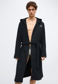 Robe de banho preta feita de tecido macio, com capuz, mangas compridas, abertura frontal com cinto na cintura e bolsos laterais. Logótipo da marca no peito.