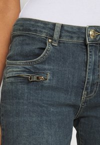 Gros plan sur la taille d'un jean bleu foncé avec bouton, braguette zippée, poche avant et petite poche zippée horizontale sur le côté gauche, porté avec un haut blanc.