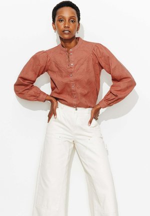 Femme debout, les mains sur les hanches, portant une blouse boutonnée texturée couleur rouille avec des manches bouffantes et un pantalon blanc taille haute à jambes larges.