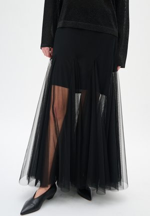Schwarzer transparenter Maxi-Rock mit fester Innenschicht oberhalb der Knie, kombiniert mit schwarzen spitzen, flachen Schuhen und einem Langarmtop.