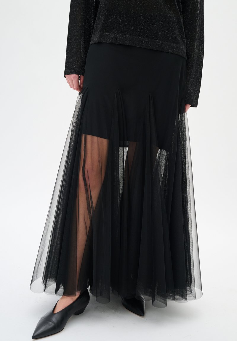 Jupe maxi noire en voile avec une couche intérieure solide au-dessus des genoux, associée à des chaussures à talons bas noires à bout pointu et un haut à manches longues.