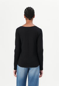 Svart langermet topp med avrundet halslinje og teksturert stoff. Modellen har på seg lyseblå vide jeans, sett fra ryggen.