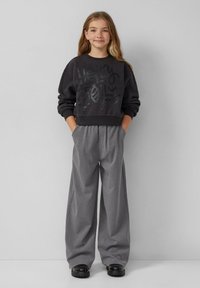 Czarny sweatshirt z graficznym nadrukiem, w połączeniu z szerokimi szarymi spodniami. Ubranie ma luźny krój i prążkowane mankiety. Czarne obuwie dopełnia całość.