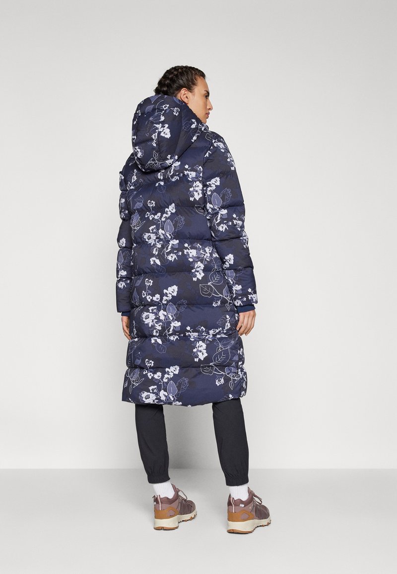 Manteau rembourré floral marin avec capuche, affichant un design matelassé et des motifs floraux blancs ; porté avec un pantalon sombre et des baskets.
