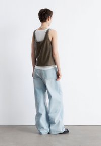 Olivgrünes Tanktop über einem weißen Shirt, kombiniert mit hellblauen weiten Jeans. Aus natürlichem Stoff, lässiges Design, sichtbare Nahtdetails.