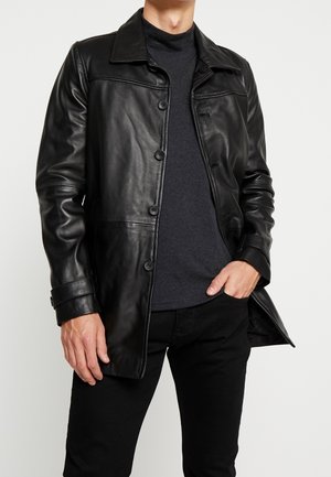 Man draagt een zwarte leren jas met knopen over een donkergrijs overhemd en zwarte broek, één hand in de zak, tegen een effen lichte achtergrond.
