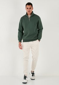 Felpa verde con zip e collo alto, abbinata a pantaloni jogger crema e sneakers in bianco e nero. Tessuto liscio e design aderente.