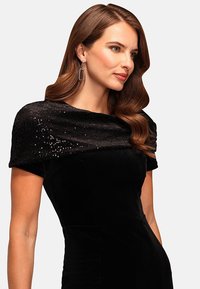Donna con capelli castani ondulati che indossa un abito in velluto nero con un colletto in paillettes e orecchini rettangolari argento, rivolta a destra.
