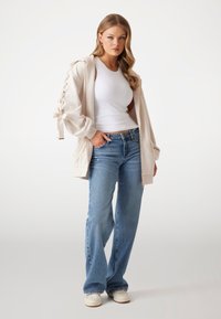 Beige hooded cardigan with lace-up sleeves, paired with a white tank top and light blue flared jeans. White sneakers complete the look.