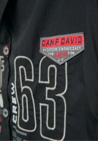 Černá látková bunda s červenými a šedými vyšívanými nášivkami, výrazným bílým prošíváním s číslem "63" a štítkem "CAMP DAVID" na hrudi.