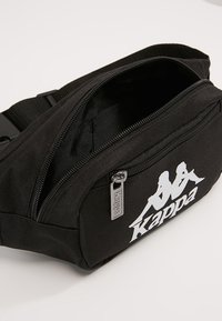 Sac banane en tissu noir avec un compartiment principal zippé, arborant un logo Kappa blanc à l'avant et un détail de tirette en métal.