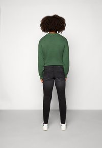 Suéter de punto verde con cuello redondo, combinado con jeans negros ajustados y zapatillas blancas. El cabello es rizado y de longitud media.