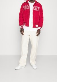 Röd puffjacka med vit "TOMMY"-text och randiga detaljer, i kombination med en vit skjorta och krämfärgade byxor; vita skor.