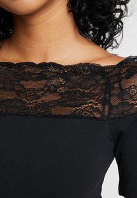 Mujer con una blusa negra de escote de encaje ancho que revela sus clavículas y cabello negro rizado parcialmente visible.
