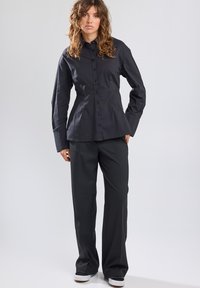 Black button-up shirt with a structured fit, long sleeves, and a curved hem. Paired with loose-fitting black trousers and white sneakers.