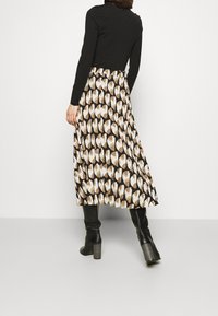 Maxirock mit plissiertem Design in einem geometrischen Muster aus Creme- und Brauntönen. Schwarzes figurbetontes Oberteil und kniehohe Stiefel mit Absatz.