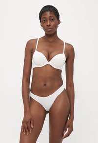 Vrouw met kort haar die een witte beha en bijpassend ondergoed draagt, staande tegen een effen lichte achtergrond.
