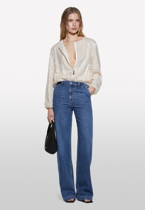 Veste crème recouverte de sequins avec un col en V profond et une fermeture éclair, associée à un jean bleu taille haute à jambes larges et un sac à main noir en tissé.