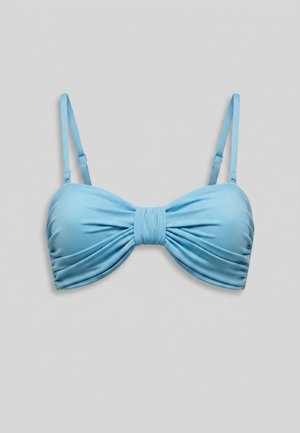 Mujer de pie descalza con bikini azul claro con top anudado y bragas fruncidas sobre un fondo liso y claro.