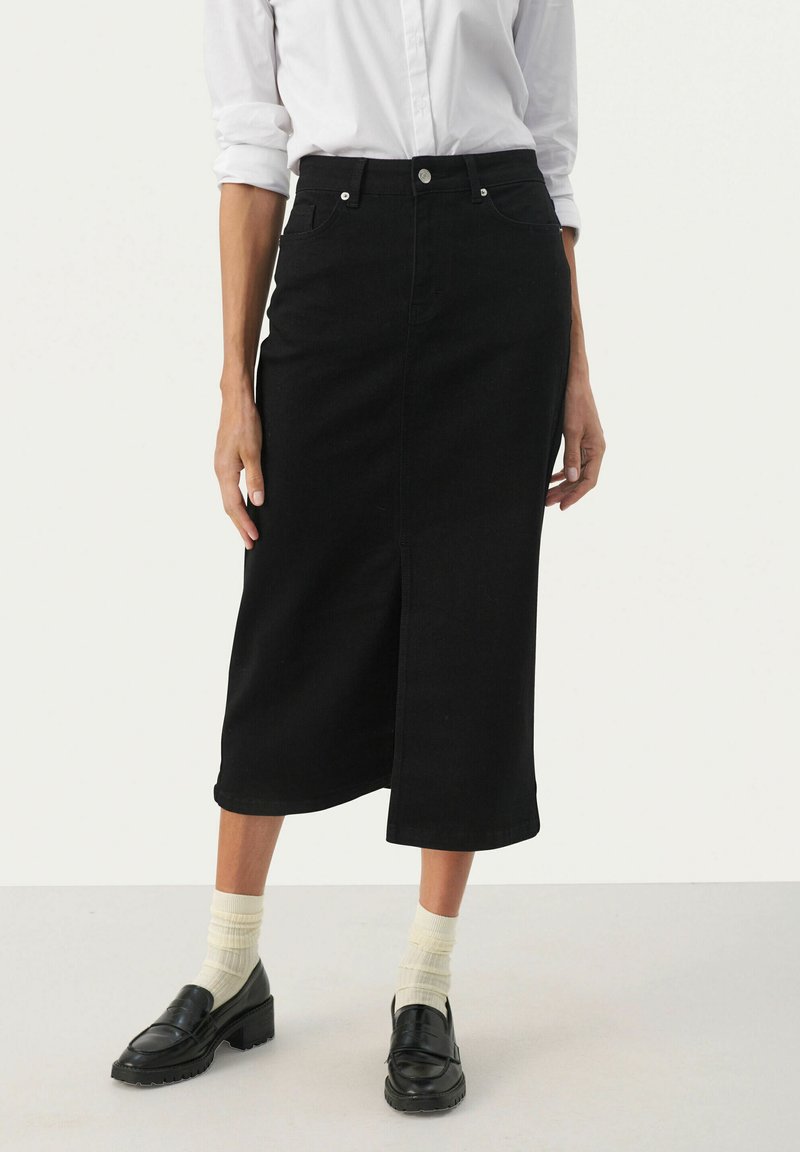 Svart denim midi kjol med en framfickslits, metallknappstängning och traditionell femfickedesign. Matchad med svarta skor och ljusa strumpor.