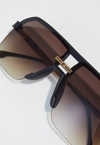 Ochelari de soare supradimensionați negri, cu lentile gradient maro, având un design elegant și un accent metalic auriu la pod. Brațe texturate.