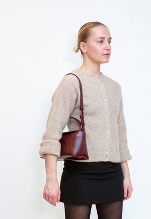 Frau mit blondem Haar im Dutt, trägt beige Strickjacke und schwarzen Rock, trägt eine braune Lederschultertasche und steht vor weißem Hintergrund.