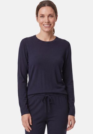 Frau mit braunem, zurückgebundenem Haar, die ein dunkelblaues Langarmshirt und passende Hose mit Kordelzug trägt, lächelt und schaut nach vorne.