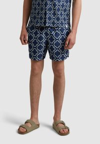 Shorts de bain navy avec un motif géométrique blanc, fabriqués en tissu léger. Associés à des sandales olive avec des sangles réglables.