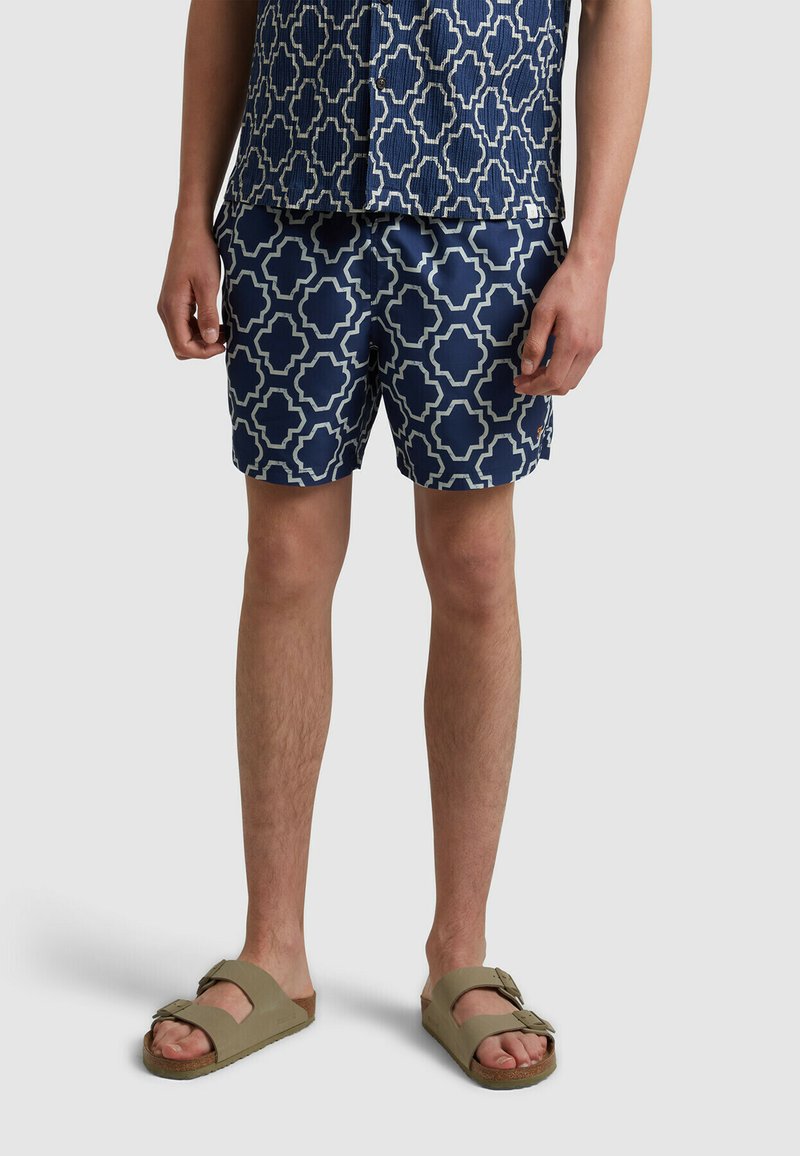 Shorts de bain navy avec un motif géométrique blanc, fabriqués en tissu léger. Associés à des sandales olive avec des sangles réglables.