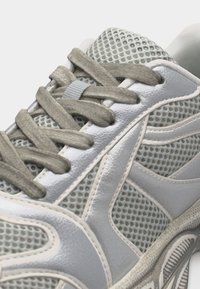 Gros plan sur une chaussure de sport grise et blanche en tissu mesh avec des lacets épais noués en motif croisé.
