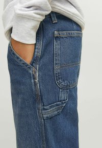 Traperice slim fit, s tamnim plavim materijalom, vidljivim šavovima, bočnim džepom i mekanom teksturom.
