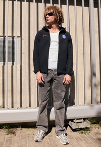 Sort zip-up hættetrøje med rødt Nike-logo, hvid tanktop, grå denim jeans og grå sneakers med accenter. Modellen har krøllet hår og solbriller.