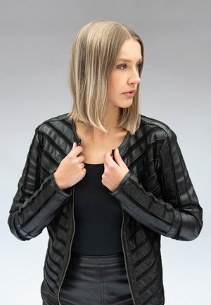 Donna bionda che si sistema una giacca di pelle nera, indossa un top nero e una gonna, guarda verso destra su uno sfondo grigio neutro.