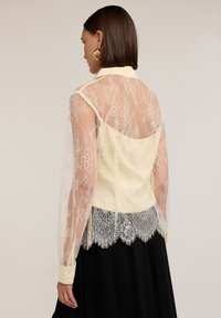Kanten blouse met een gele kraag en lijfje, doorzichtige lange mouwen en een gescandeerde kanten zoom. Bevat een gedetailleerd bloemenpatroon.