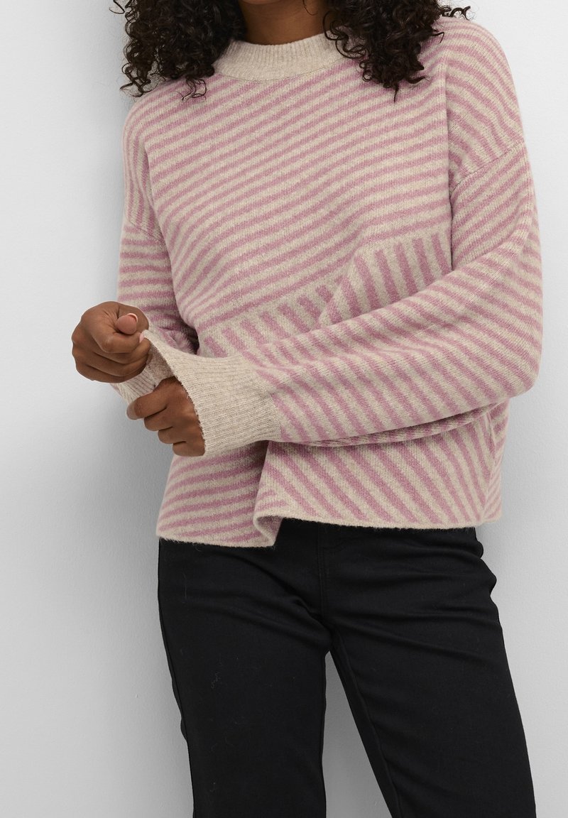 Femme portant un pull rayé rose et beige avec des poignets côtelés, ajustant une manche, associée à un pantalon noir devant un fond uni.