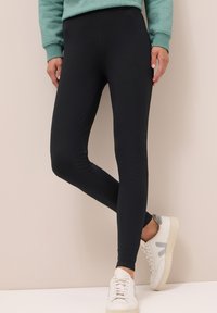 Leggings noirs en matériau extensible, présentant un design épuré et une taille haute, associés à des baskets blanches pour le contraste.