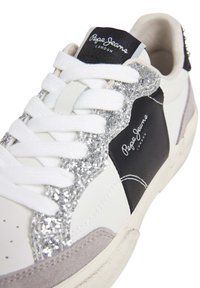 Sneaker bianca con accenti di glitter neri e argenti. Presenta un pannello in suede grigio testurizzato, lacci bianchi e una toppa nera con logo sulla linguetta.