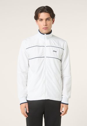 Ung mand med mørkt hår iført en hvid FILA lynlåstrøje med marineblå kant og sorte bukser, stående foran en ensfarvet lys baggrund.