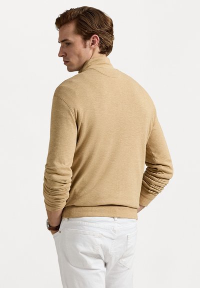 Šviesiai ruda golfo tipo megztinis su minkšta tekstūra, ilgomis rankovėmis ir prigludusia forma, derinamas su baltais kelnais. Matomas prekės ženklas nėra.