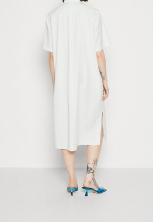 Abito camicia bianco a maniche corte con piega sul retro, dotato di fessure laterali. Abbinato a mule blu lucidi e mette in mostra tatuaggi sulle gambe.