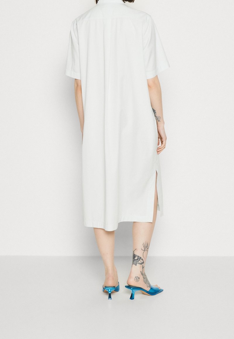 Abito camicia bianco a maniche corte con piega sul retro, dotato di fessure laterali. Abbinato a mule blu lucidi e mette in mostra tatuaggi sulle gambe.