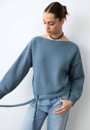 Donna con capelli scuri raccolti, indossa un maglione blu largo con dettaglio al collo e jeans azzurri, in piedi davanti a uno sfondo bianco.