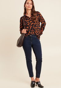 Frau steht mit einer Hand in der Tasche, trägt eine orange und schwarze Leopardenmuster-Bluse, dunkle Jeans, schwarze High-Heel-Mules und hält eine braune Umhängetasche.