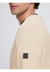 Világos bézs pulóver, texturált kötött anyagból, kerek nyakkal és sötét tengerészkék díszítéssel a gallérnál. A ujján egy apró fekete logópajzs található.