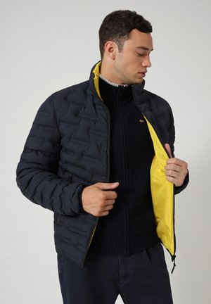 Giacca a vento nera trapuntata con interno giallo, chiusura con zip e colletto alto, indossata su un maglione blu navy. Presenta una trama liscia e un design aderente.