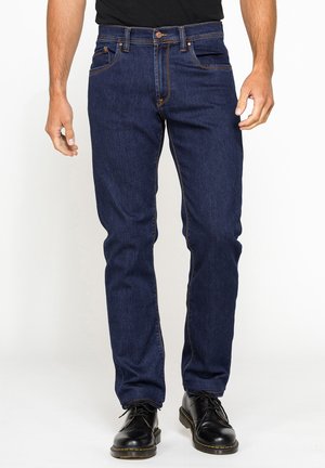 Uomo che indossa jeans blu scuro a gamba dritta e scarpe nere con lacci, in piedi su uno sfondo bianco uniforme.