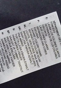 Etichetta di cura su tessuto, sfondo bianco con testo nero in più lingue. Istruzioni sul lavaggio e sulla separazione dei colori.