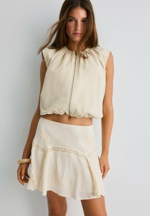 Jeune femme portant un haut beige sans manches zippé et une jupe crème avec broderies florales, posant devant un fond gris uni.