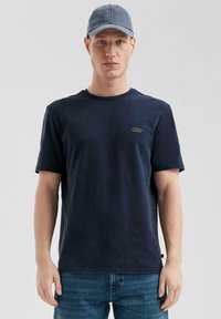 Jonge man met een marineblauwe pet, een donkerblauw T-shirt met kleine tekst op de linkerkant van de borst en blauwe spijkerbroek, staand tegen een effen lichte achtergrond.